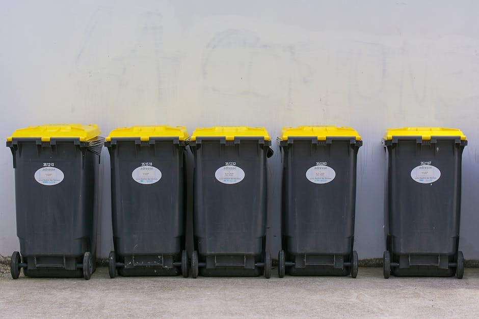Five large black rubbish bins with yellow lids are lined up on a concrete surface against a plain, light-colored wall. The bins are made of durable plastic with textured surfaces, featuring small whit