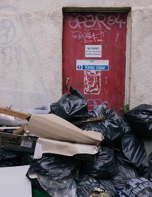 Three large black plastic rubbish bags, filled and tied at the top, are stacked along the edge of a paved sidewalk next to a curb. The bags appear to contain household waste or yard debris and are pos